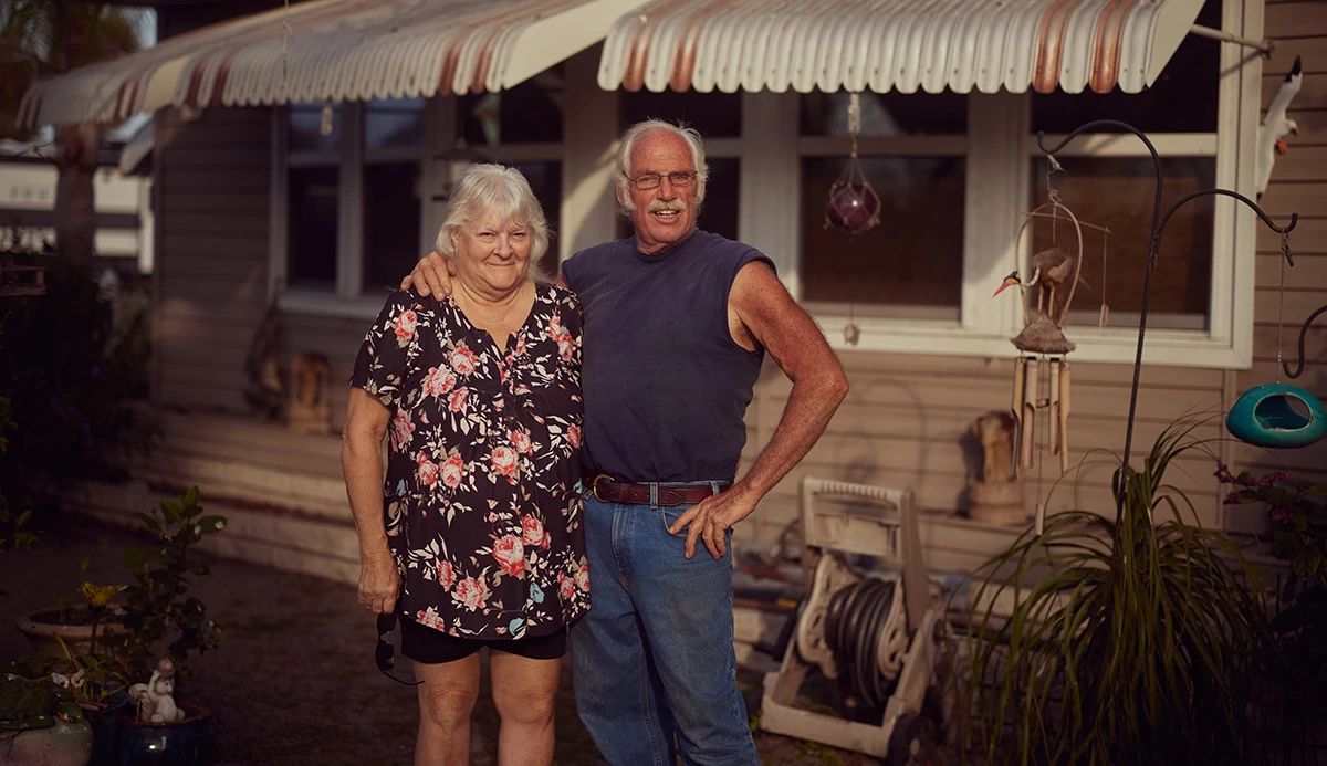 Woman in flowered shirt & man in sleeveless T-shirt in front of mobile home