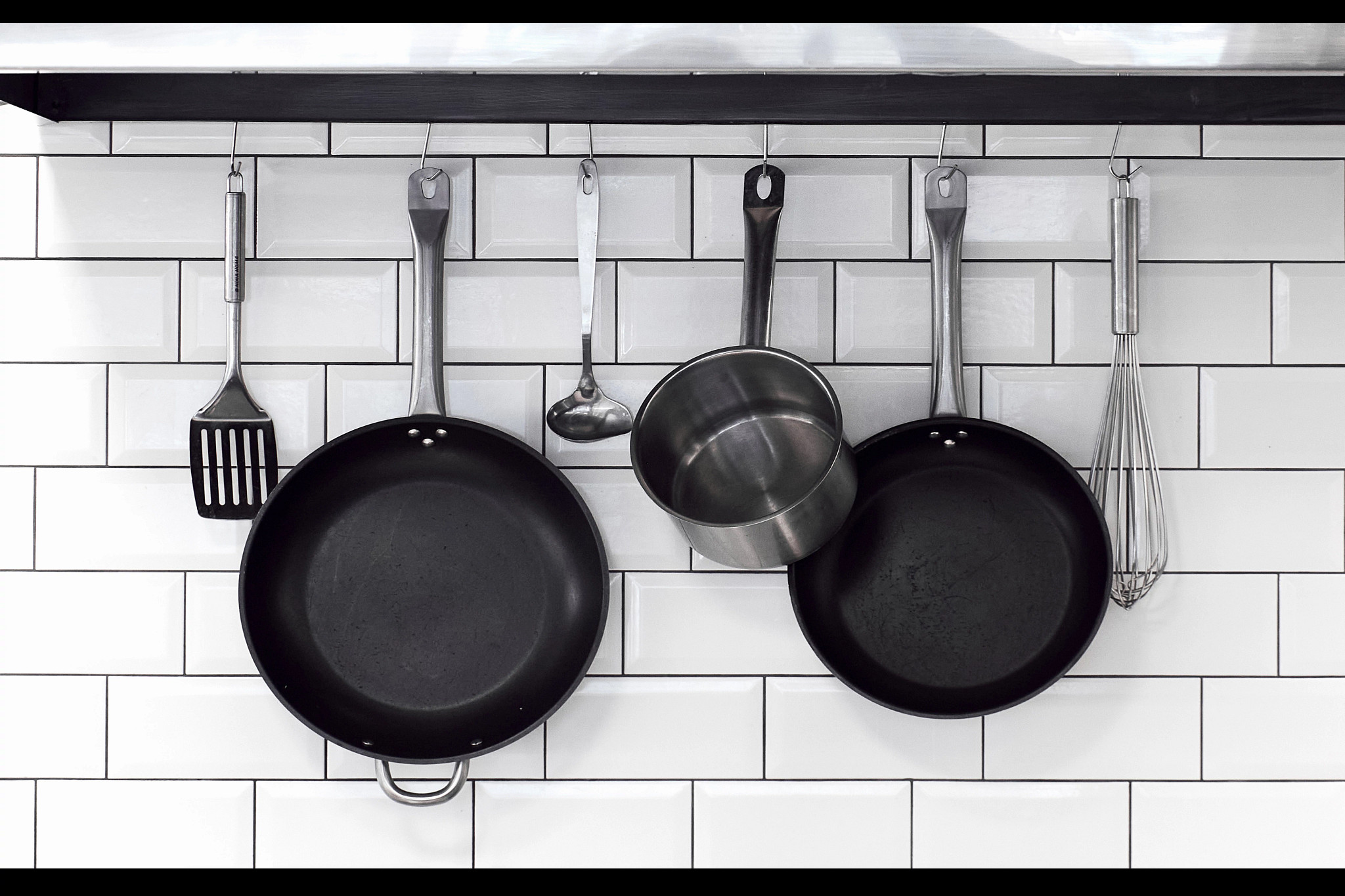 scratched nonstick pans, stainless steel utensils and a stainless steel pot are shown hanging on a tile wall in a kitchen