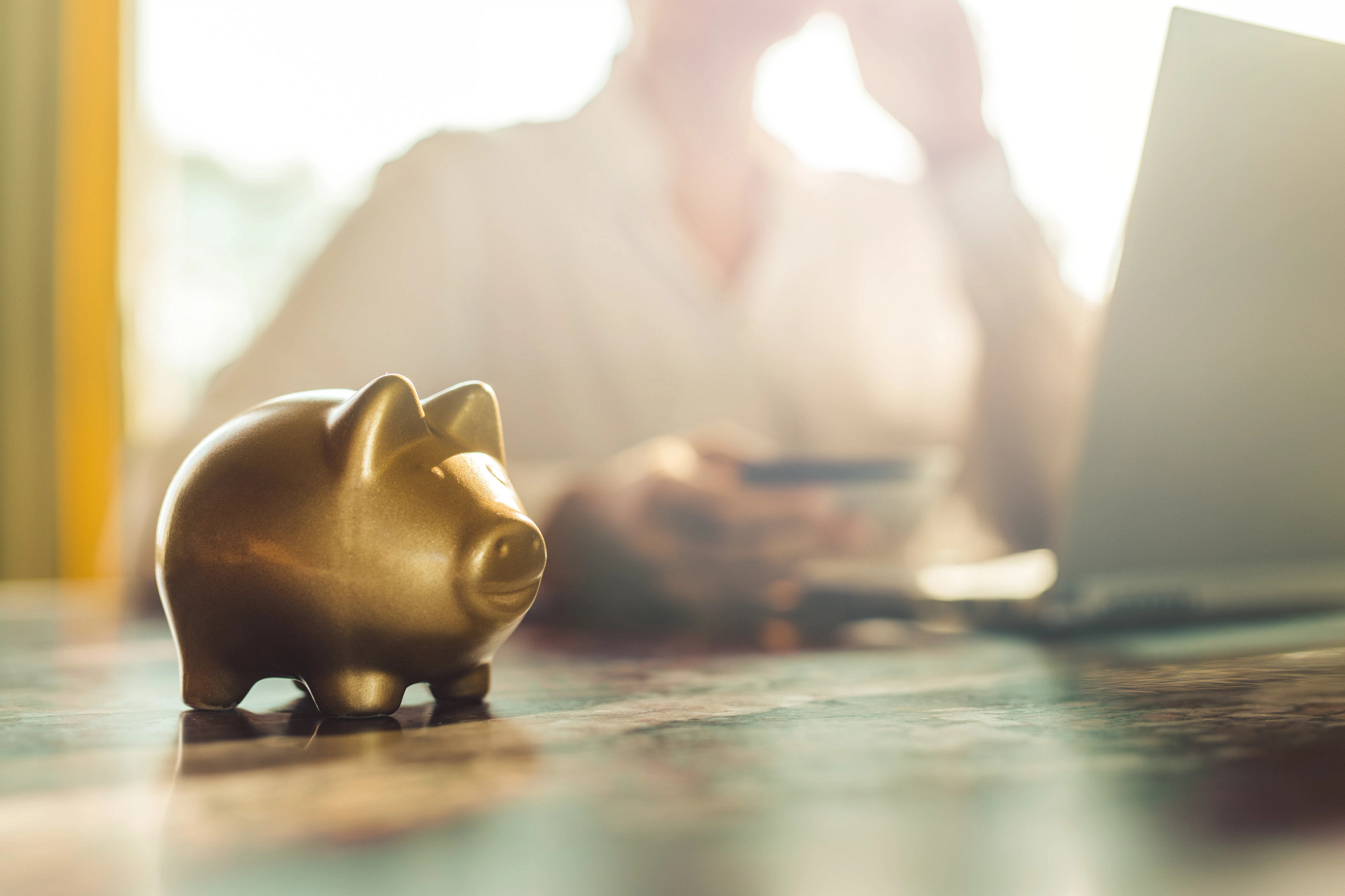  A gold piggy bank sits on a wooden desk, illuminated by natural sunlight streaming through a window.