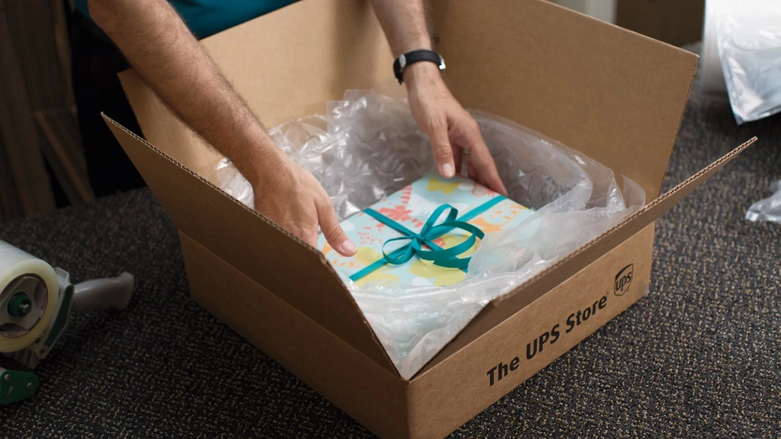 hands putting a birthday wrapped gift box in a U P S shipping box