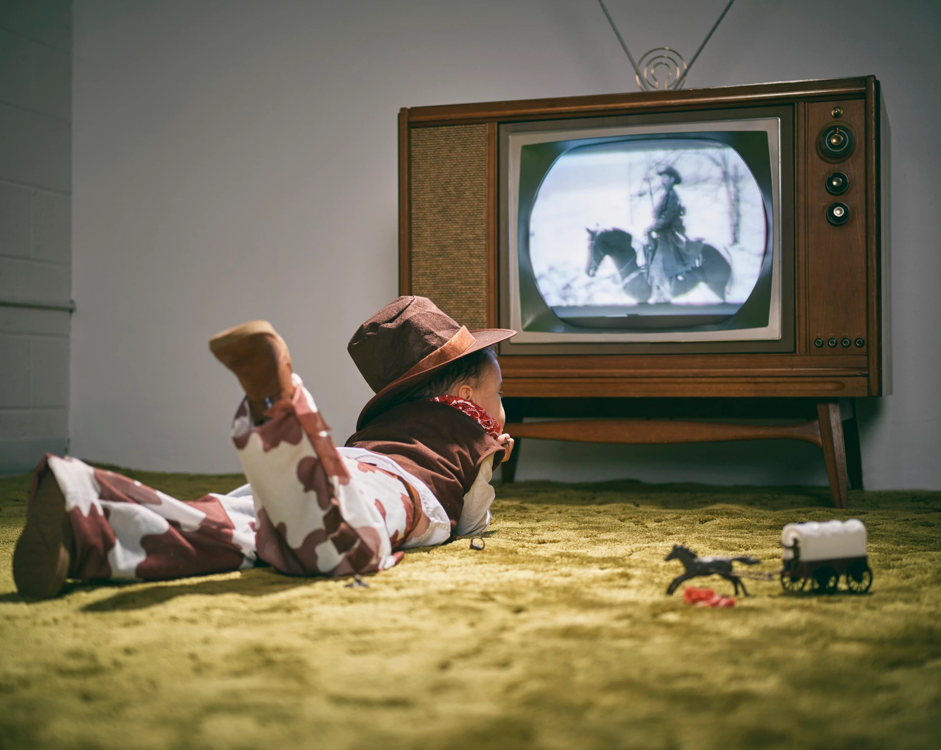 A period correct 1960's television displaying a cowboy scene on the screen with a little boy dressed as a cowboy watching the screen.