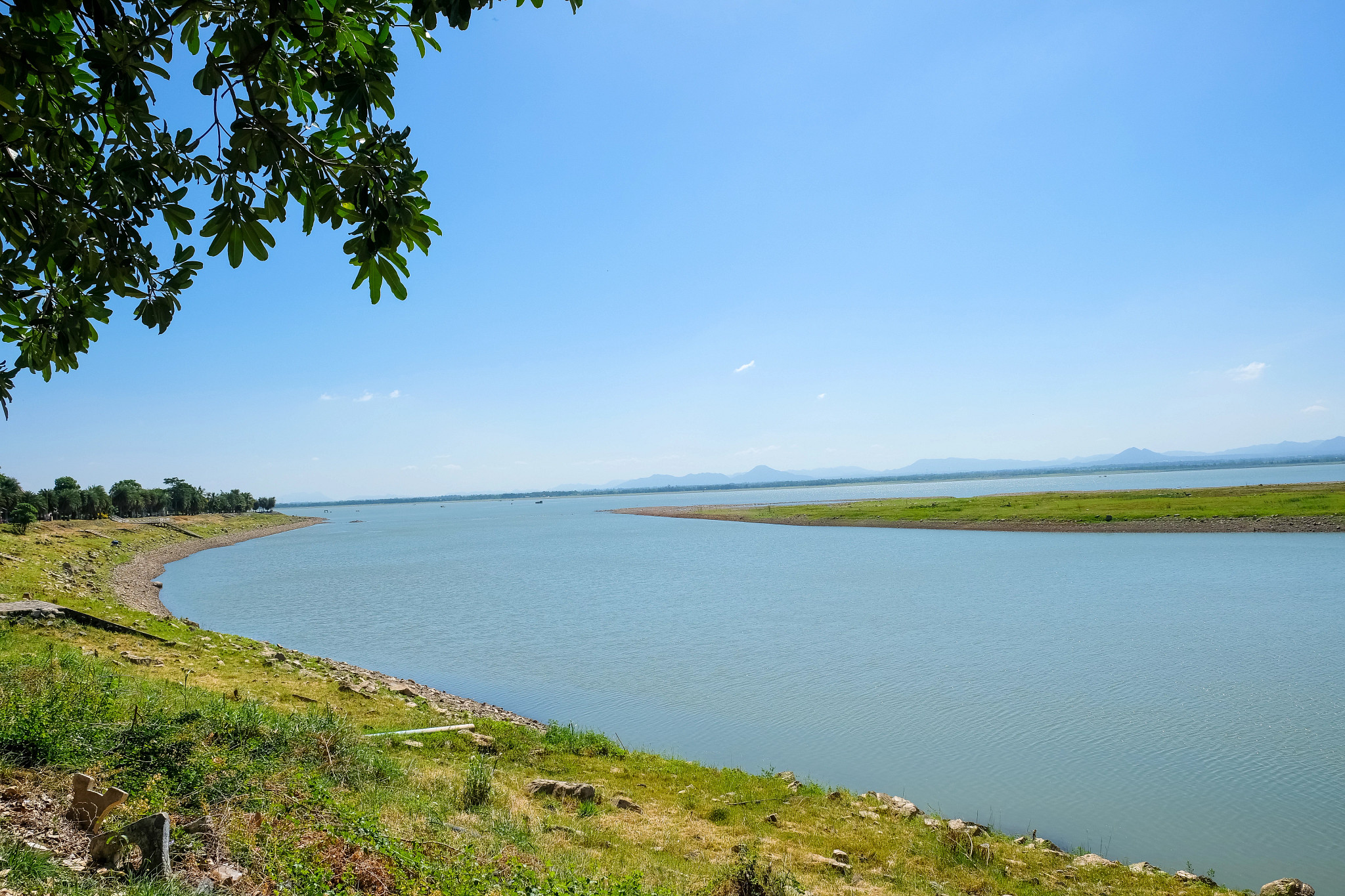 Calm lakeside landscape with clear blue sky, tree branches framing the view, and distant hills across the water