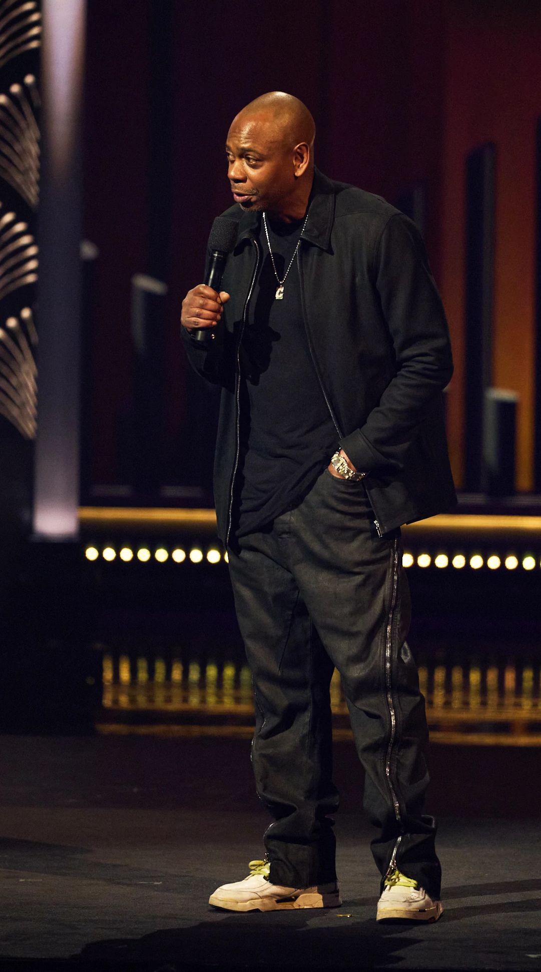 Dave Chappelle speaking into a microphone onstage at the 25th Annual Mark Twain Prize For American Humor