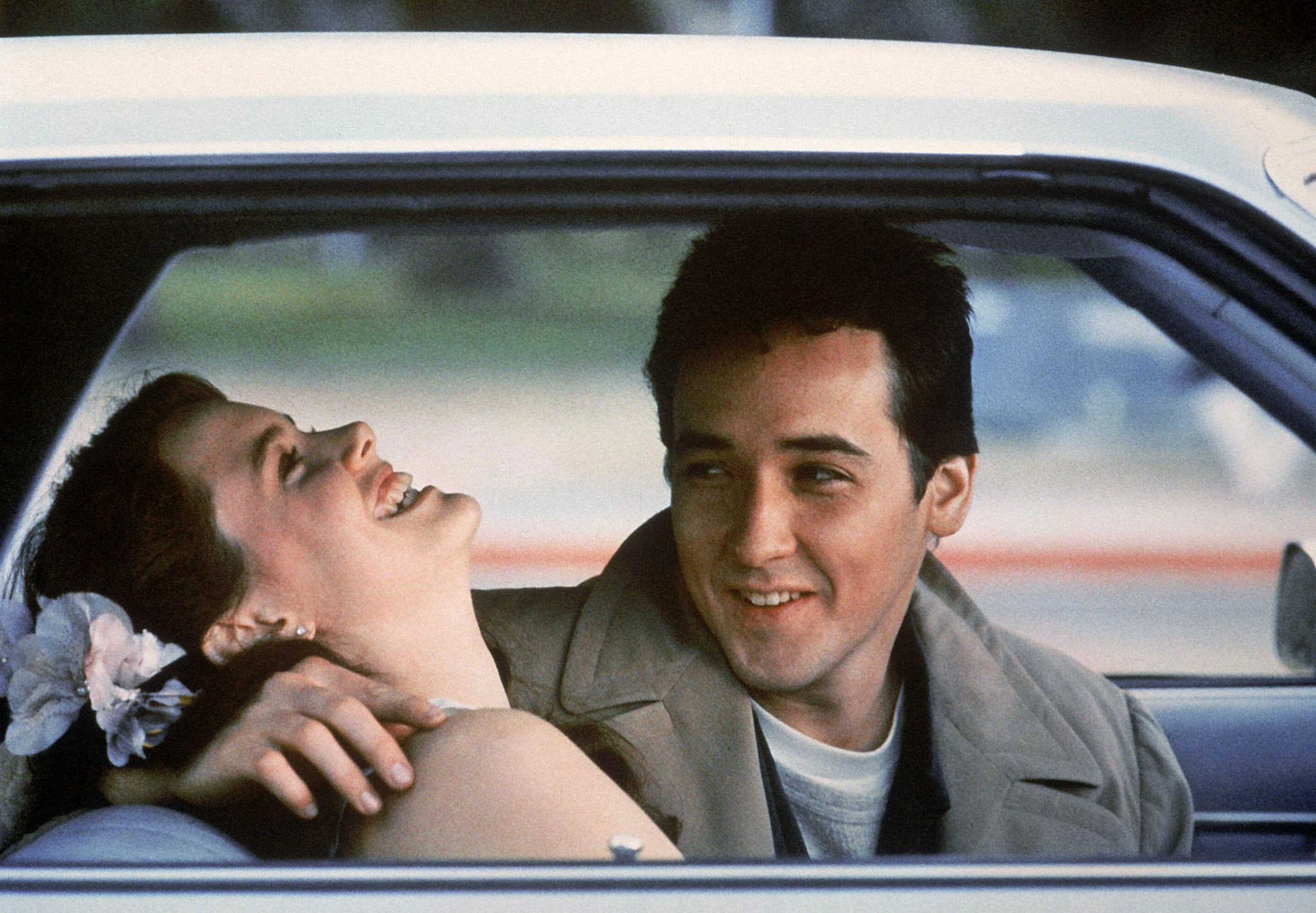Ione Skye and John Cusack seated in a car lean close while sharing a lighthearted moment in the film Say Anything