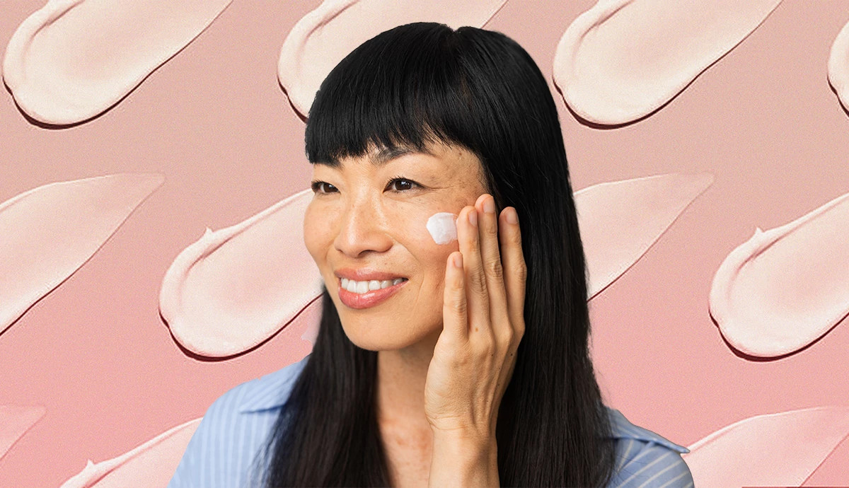 Smiling woman applying white face cream to her cheek against a pink patterned background