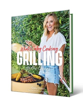 Cookbook ‘What’s Gaby Cooking: Grilling All the Things’ by Gaby Dalkin Cookbook that says What’s Gaby Cooking Grilling All the Things, Gaby Dalkin; Gaby Dalkin holding lid above grill with food on it on cover