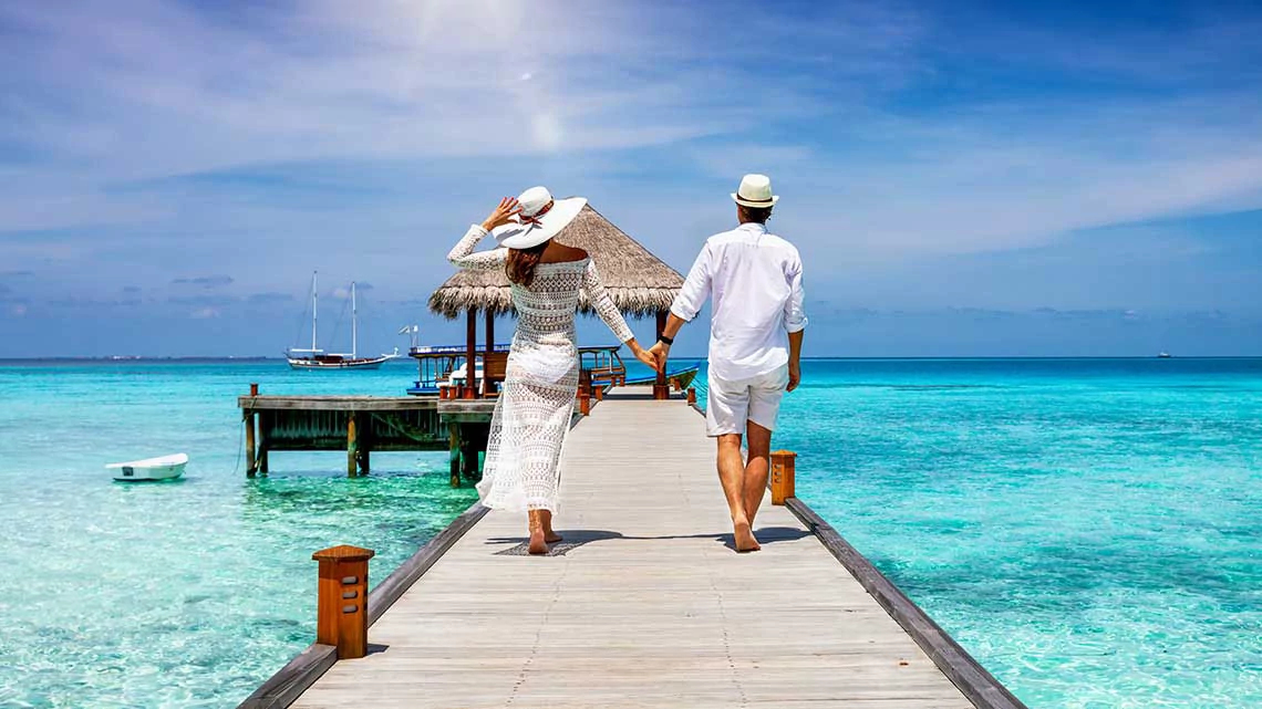 Vacation Package Savings, an AARP Member Benefit A happy couple in white summer clothing on vacation walks along a wooden pier over tropical, turquoise ocean in the Maldives, Indian Ocean