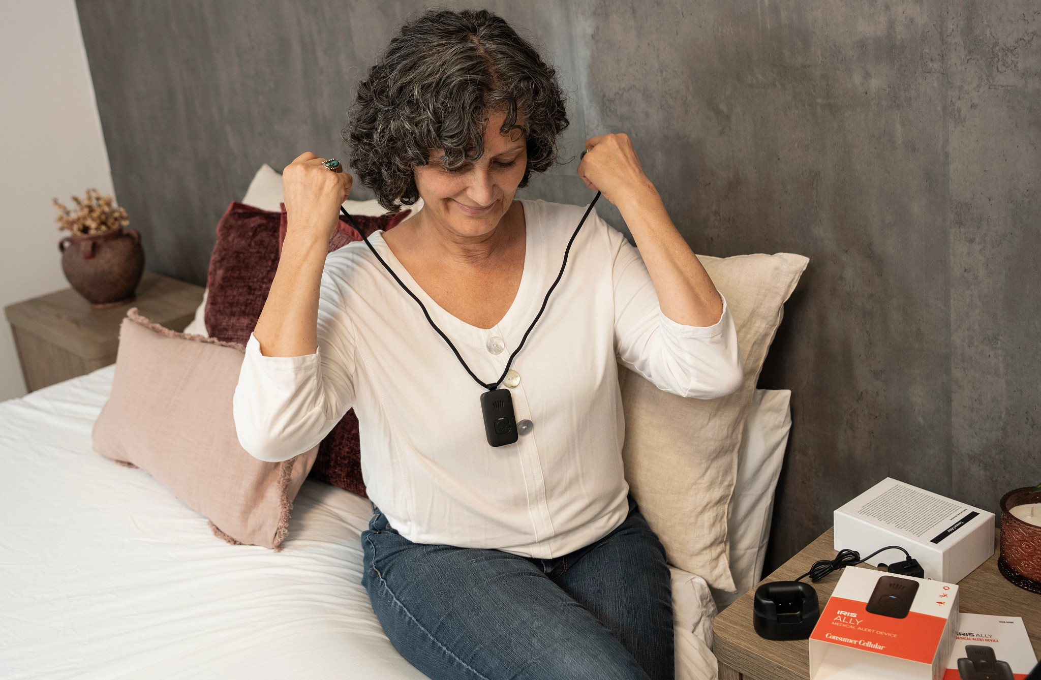A woman putting the IRIS Ally medical alert device lanyard on her neck in a bedroom