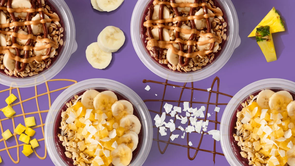 Jamba banana pieces, mango pieces, purple background, peanut butter, chocolate sauce, nuts, shaved coconut in bowls