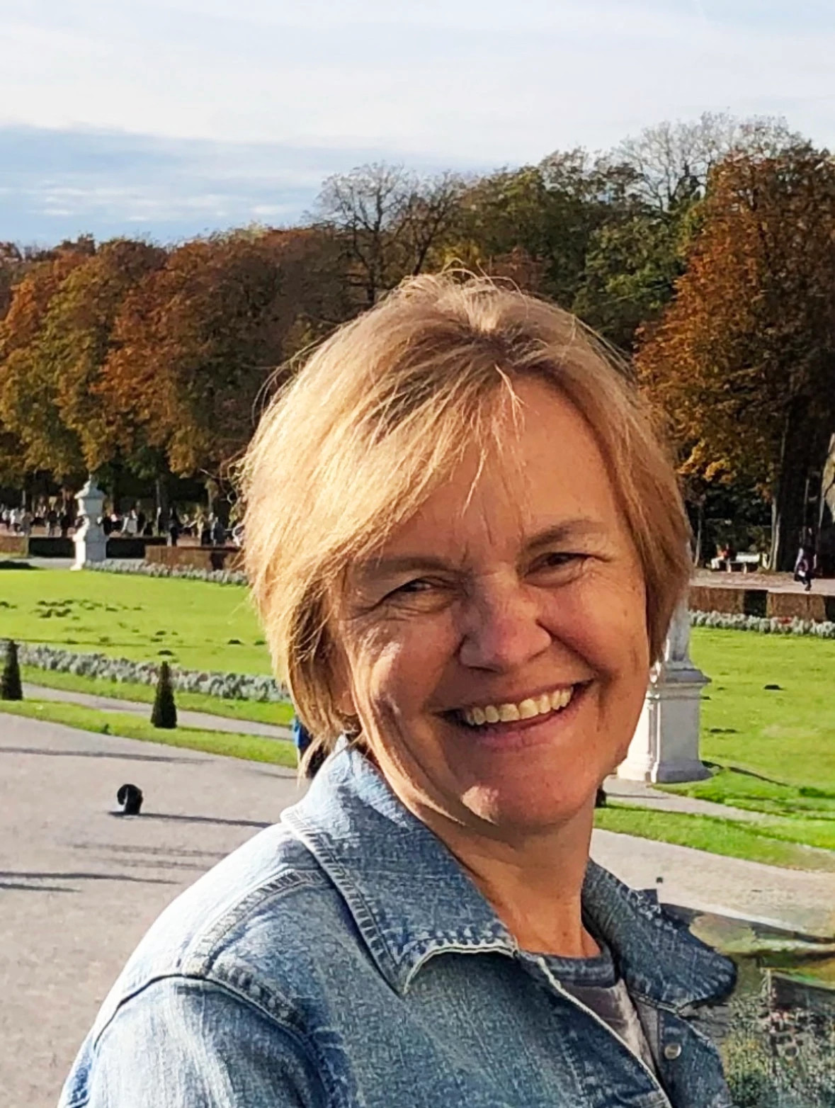 Outdoor portrait of an older adult standing in a park with green lawns and autumn trees in the background