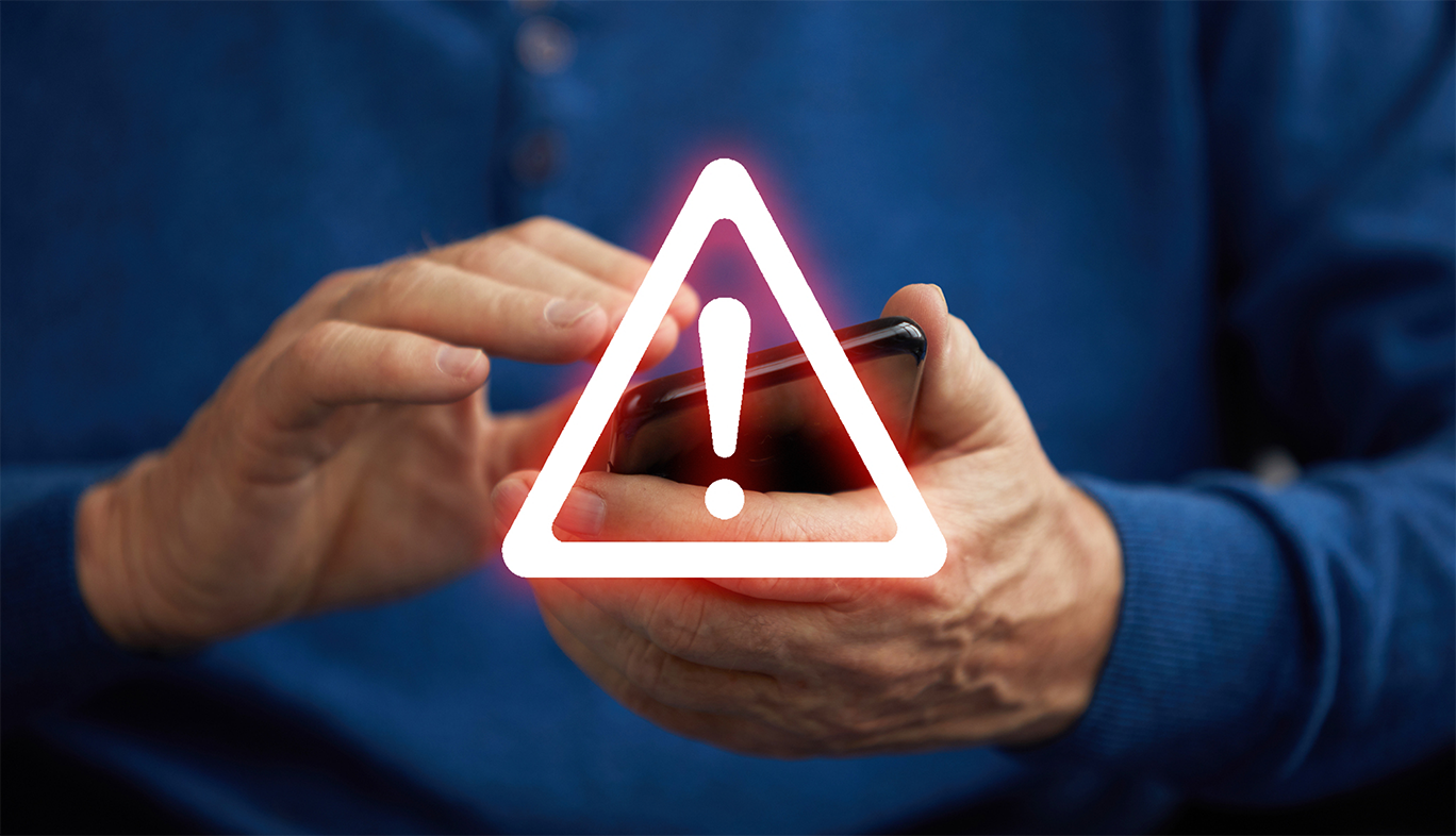 Los delincuentes contactan a potenciales víctimas a través de sus teléfonos inteligentes para iniciar la mayoría de las estafas de inversión. Un triángulo de advertencia se enciende frente a un teléfono que está en manos de una persona