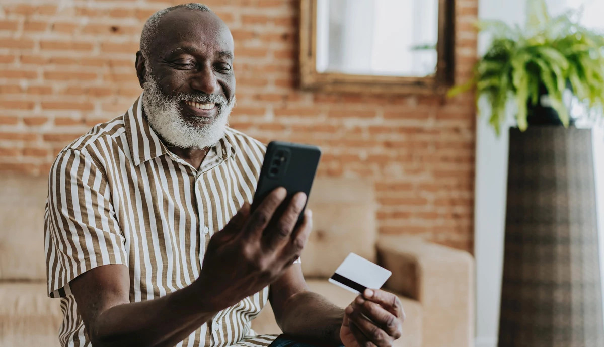 Smiling man sitting wearing striped shirt while holding cell phone and debit card in hands