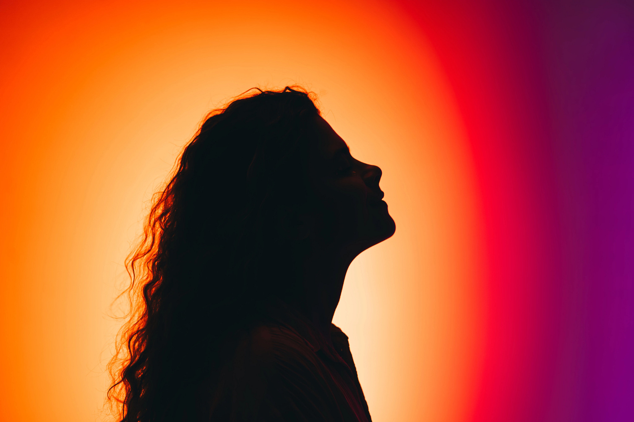 Side-profile silhouette of an adult with long wavy hair against a vivid orange, pink, and purple gradient background