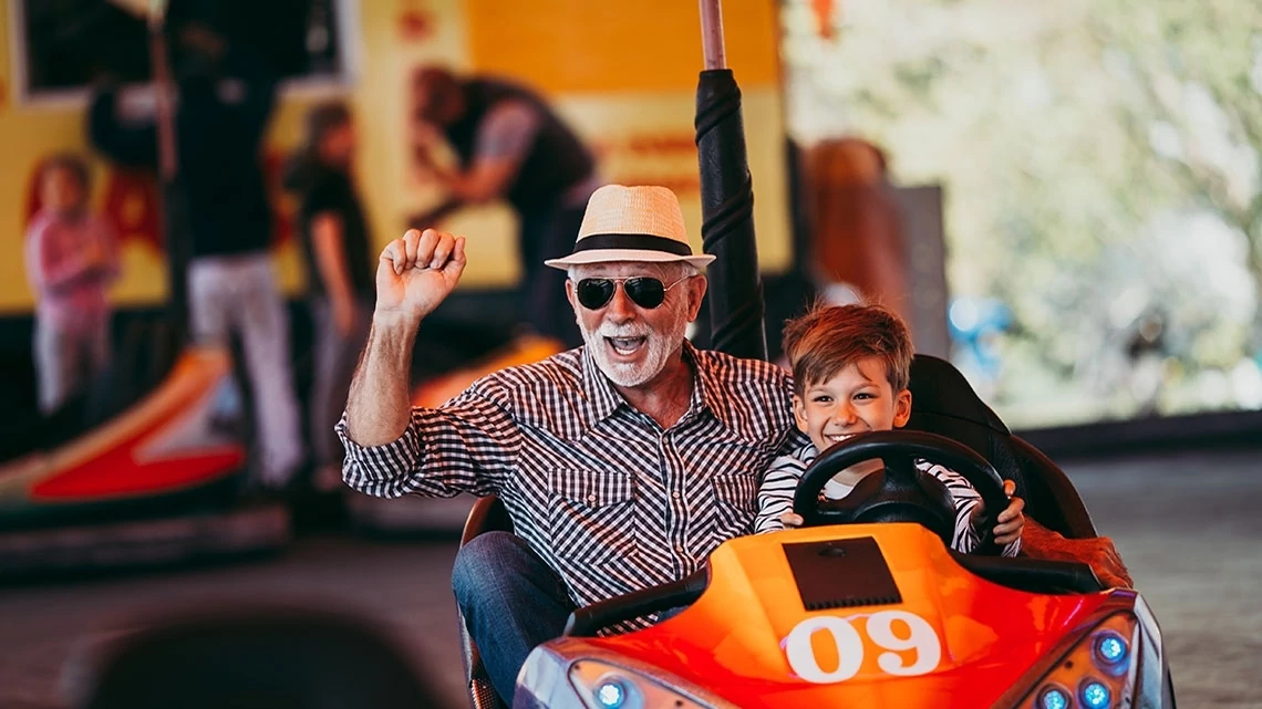 Even More Entertainment Discounts grandfather wearing sunglasses and plaid shirt with hat on and grandson riding in an orange with the number 9 on it go-cart smiling