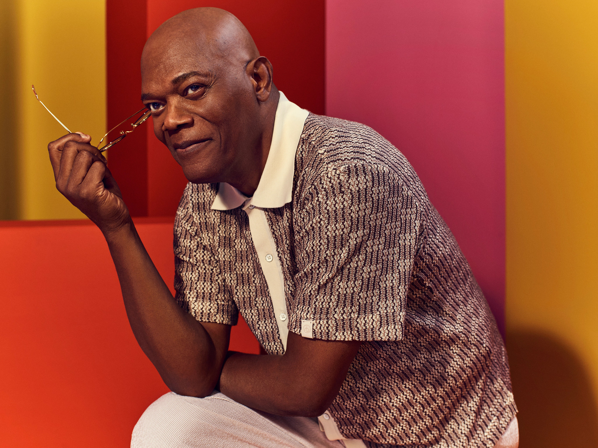 Samuel L Jackson poses for a portrait placing his elbow to lean on his knee while holding a pair of glasses in his hand