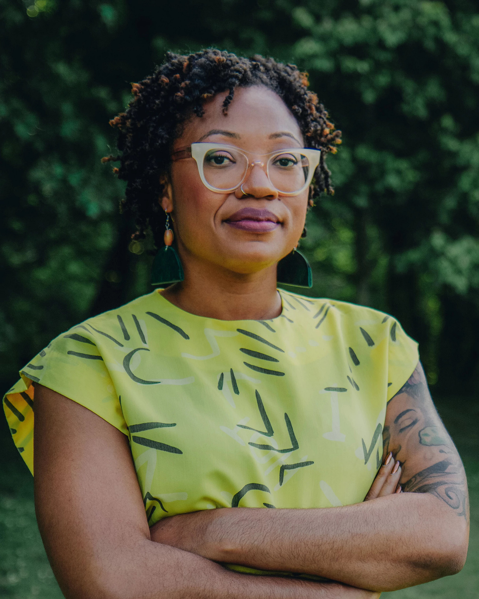 Professional outdoor headshot of Ashante Reese wearing glasses and a green patterned top with arms crossed