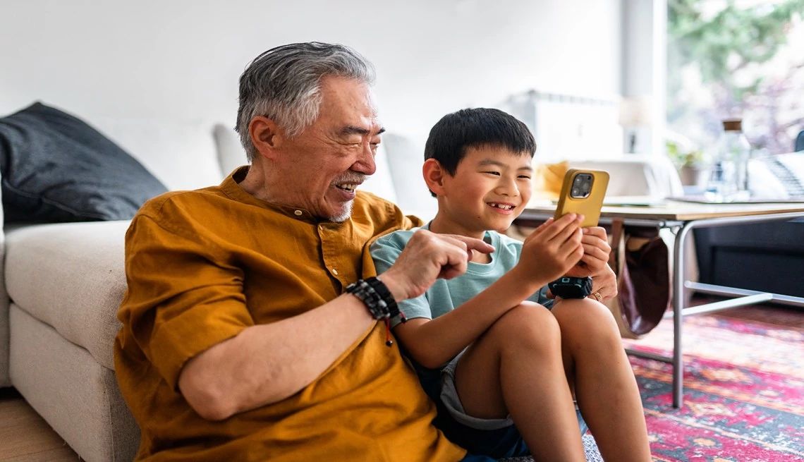 Grandfather sitting on the floor with his smiling grandson while they play a game on a smartphone together