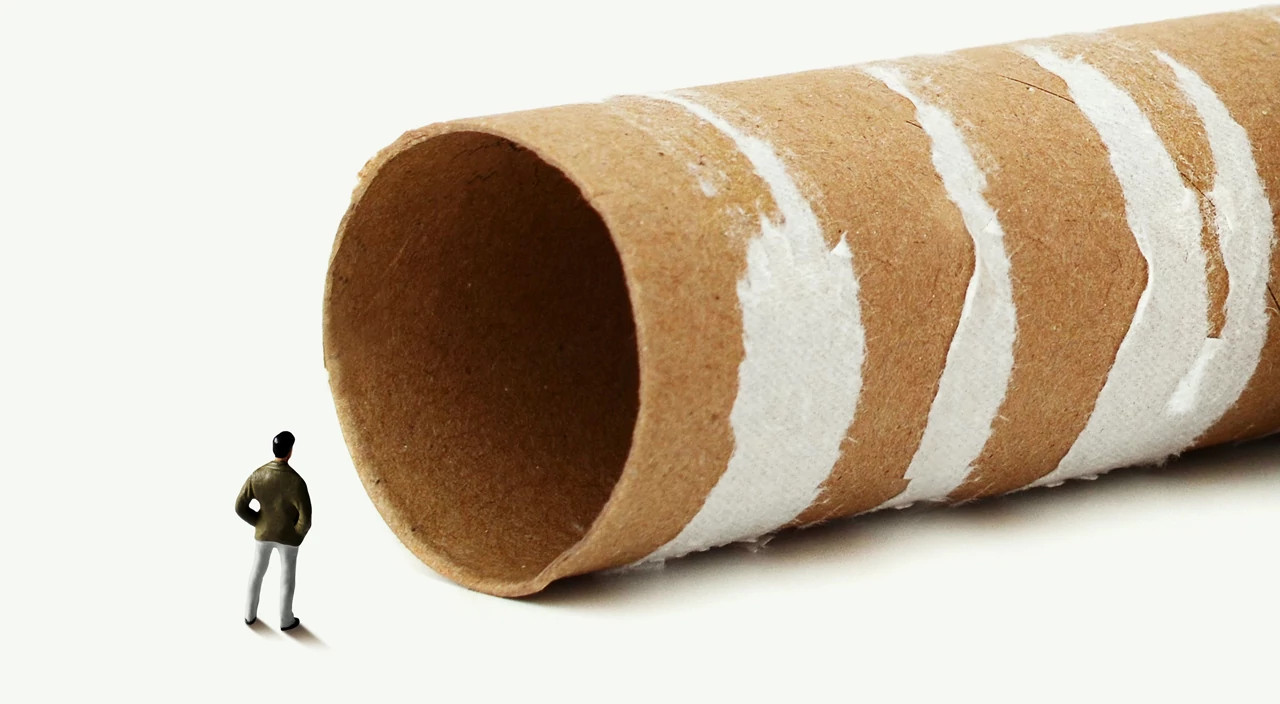 miniature toy man stands on a white surface, looking into the opening of a giant, empty cardboard toilet paper roll that looms over him, symbolizing the daunting feeling of a colonoscopy