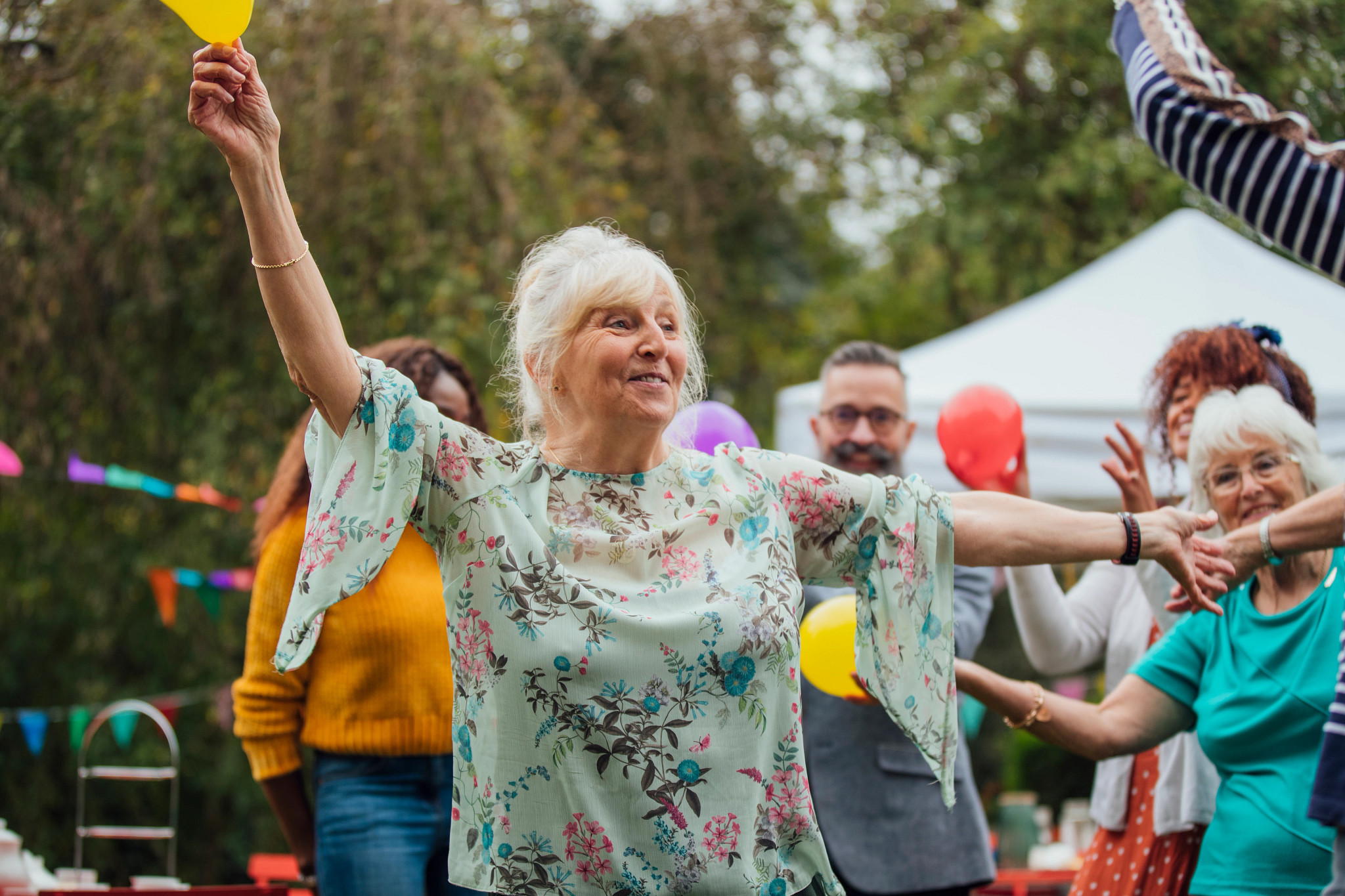 An older female enjoys dancing in the garden at an autumn tea party. Adults dance around her and colorful bunting and balloons decorate the garden.