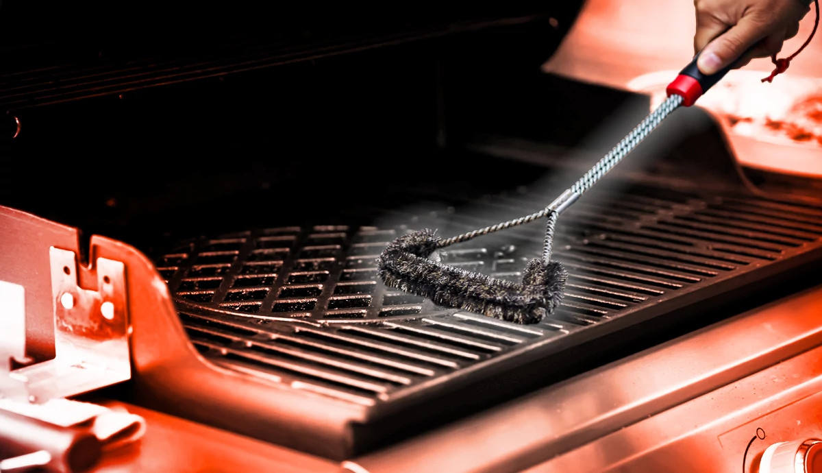 hand holds a metal wire bristle grill brush with a black and red handle, scrubbing the metal grates of a grill
