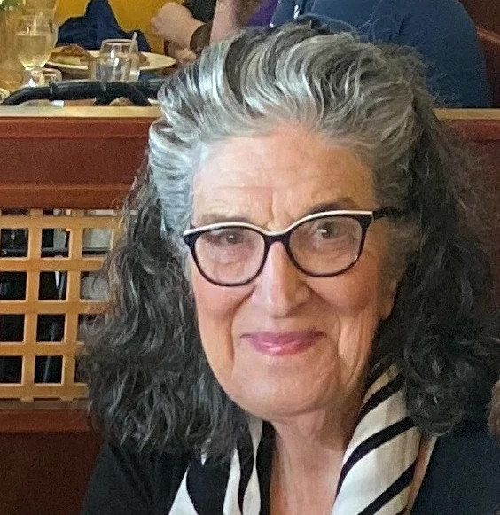 Portrait of an older adult wearing glasses and a patterned scarf while seated indoors at a restaurant