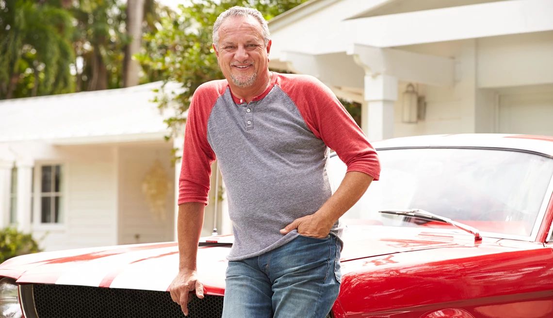 Mature man wearing blue jeans leaning on a shiny red classic car in the driveway in front of his home