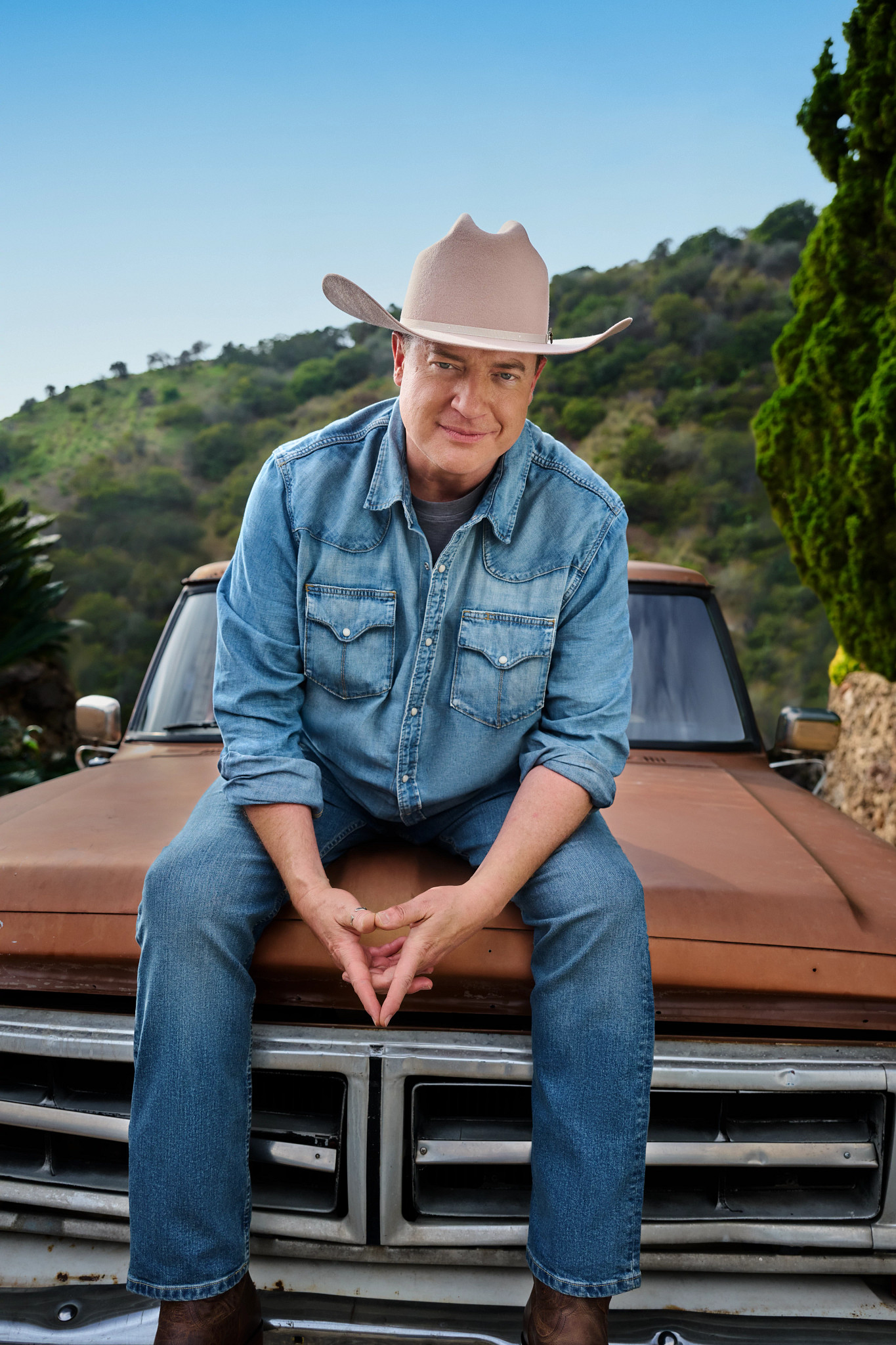brendan fraser posing for a portrait seated on the hood of a vintage pickup truck, wearing a cowboy hat and blue chambray shirt