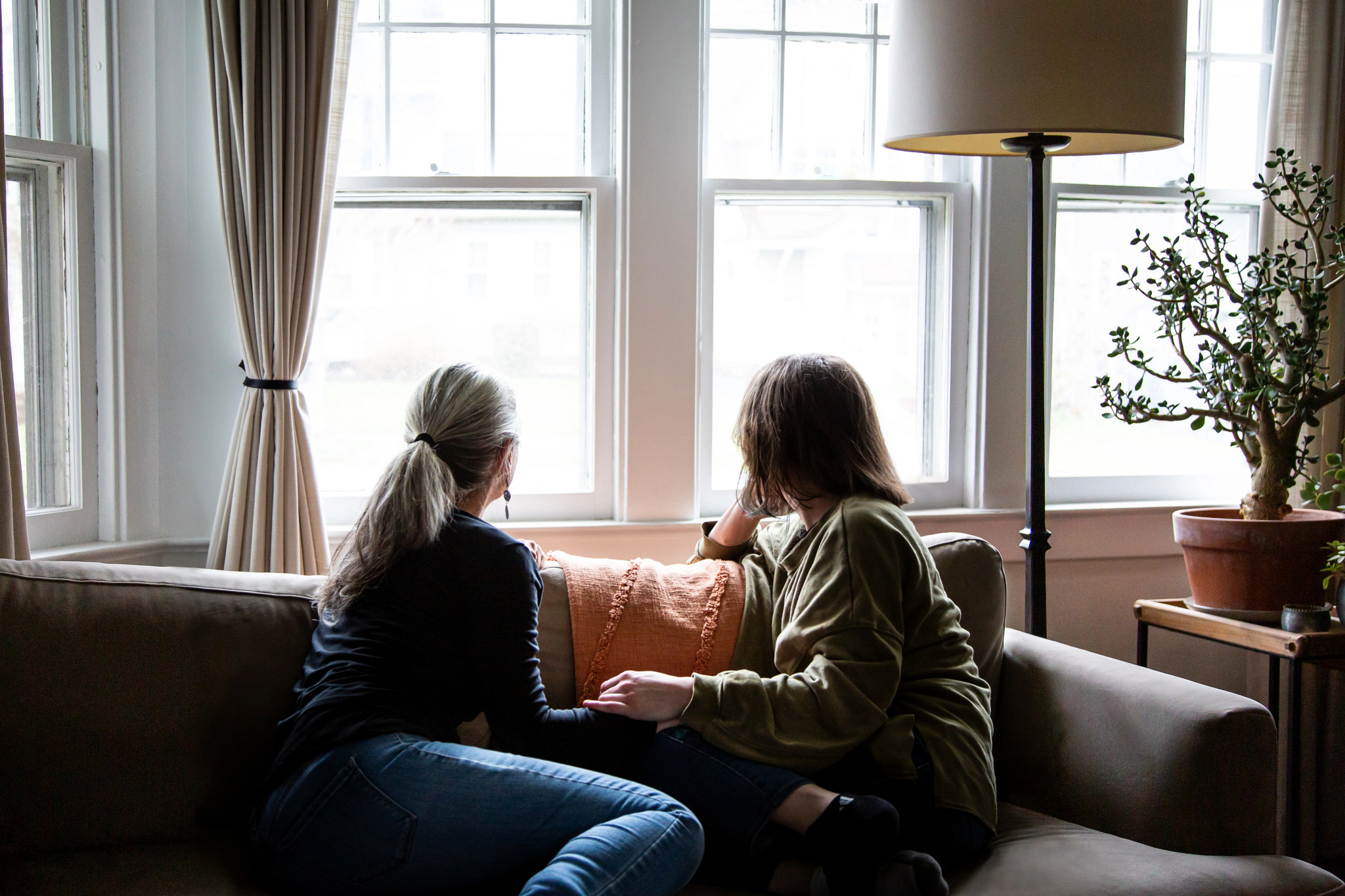 A mother and adult child sit on a couch, staring out of the windows in their home.