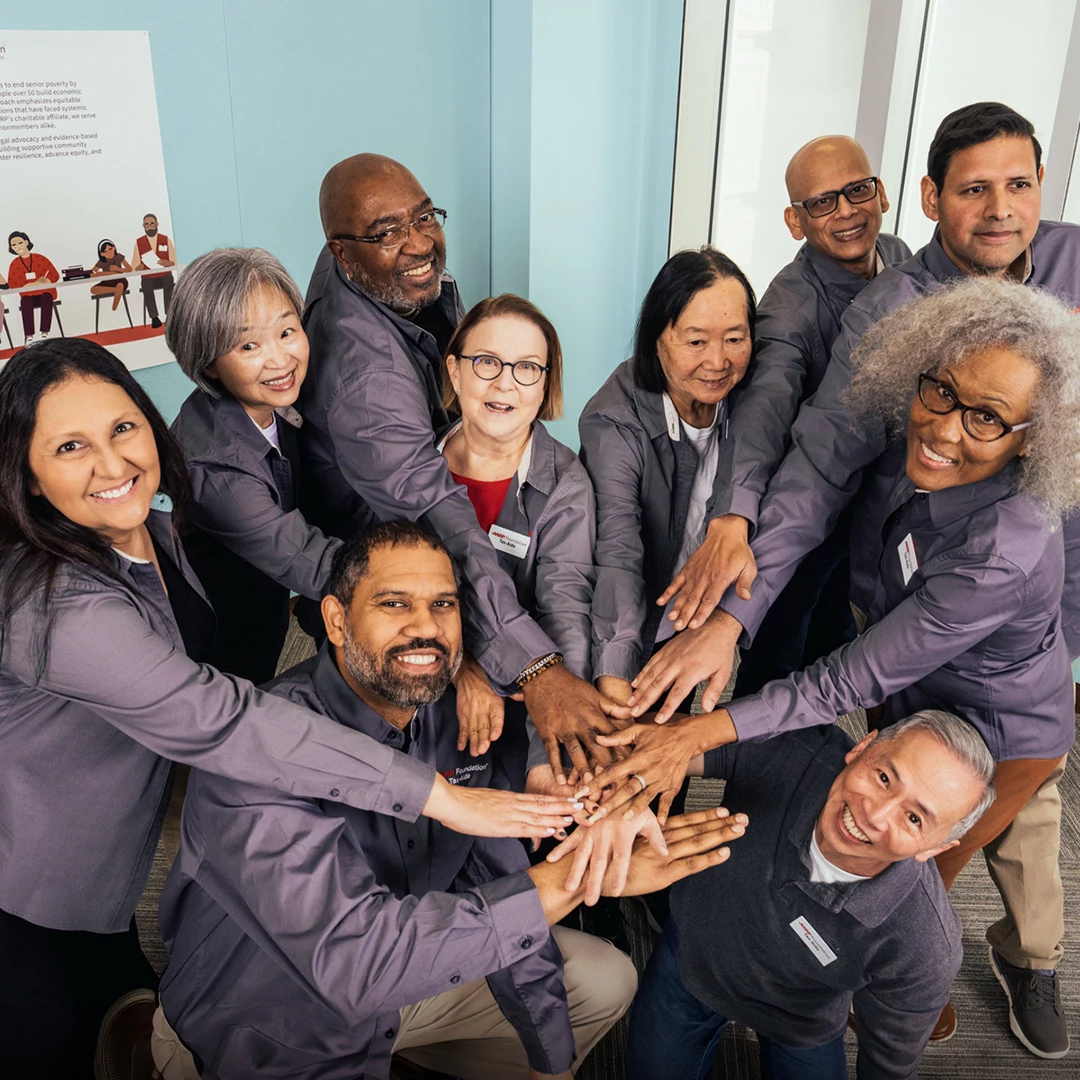 A diverse group of smiling AARP Foundation volunteers stacks their hands together in a supportive team huddle.