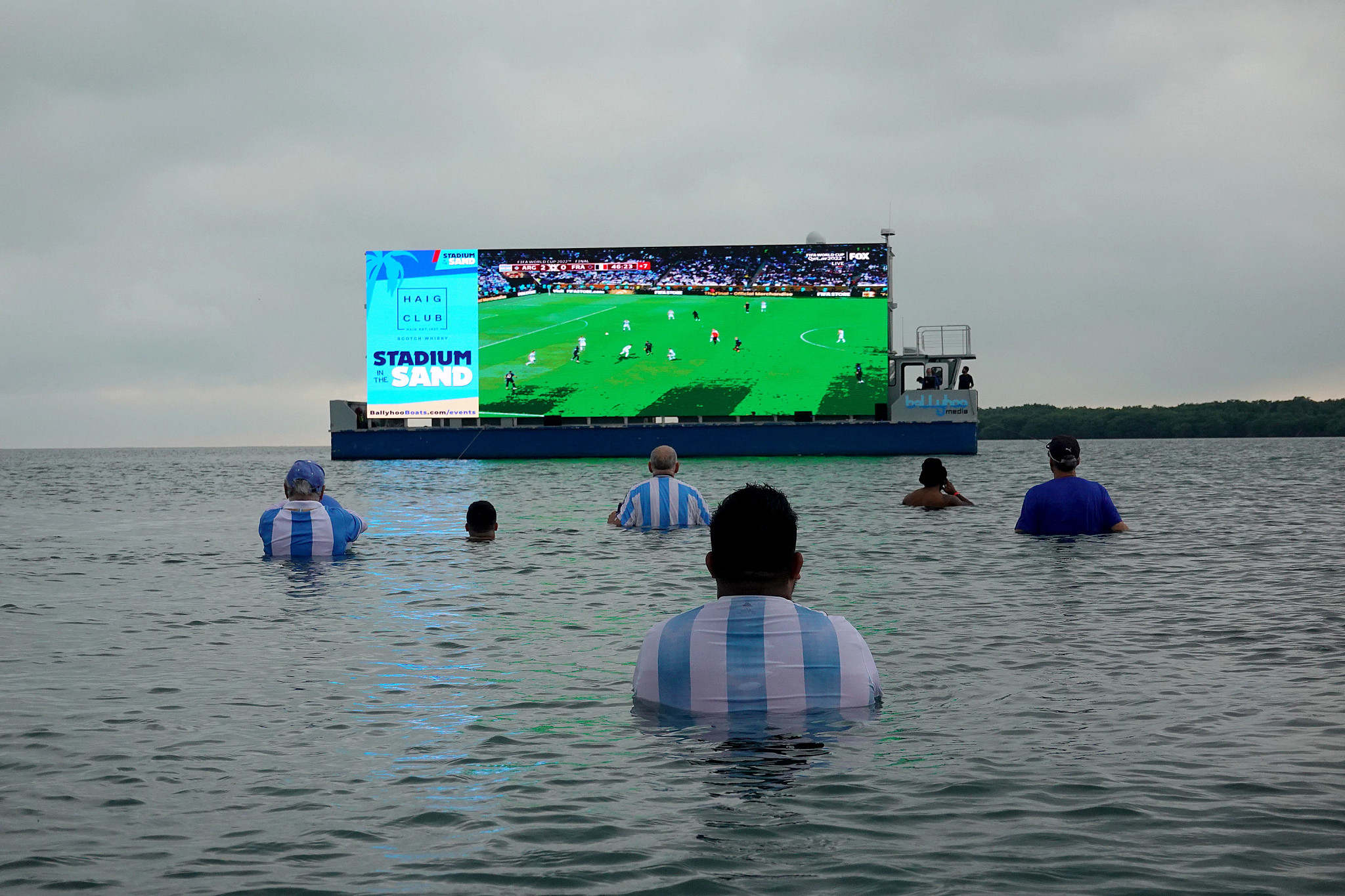 people wading in the water while watching a soccer match on a screen on a boat